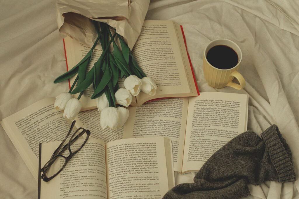 Open books, a coffee mug, white tulips, and reading glasses on a bed