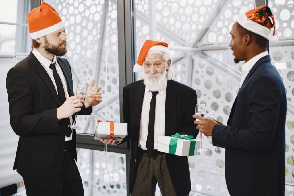 Three businessmen in Santa hats exchanging gifts and toasting at a holiday party indoors