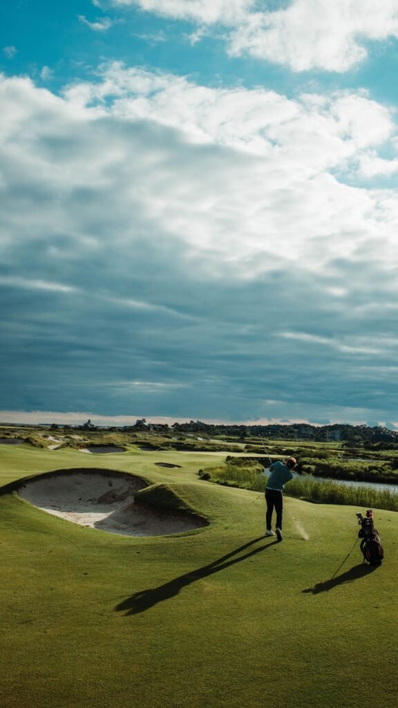 A golfer swinging on a links course beside a bunker under a cloudy sky