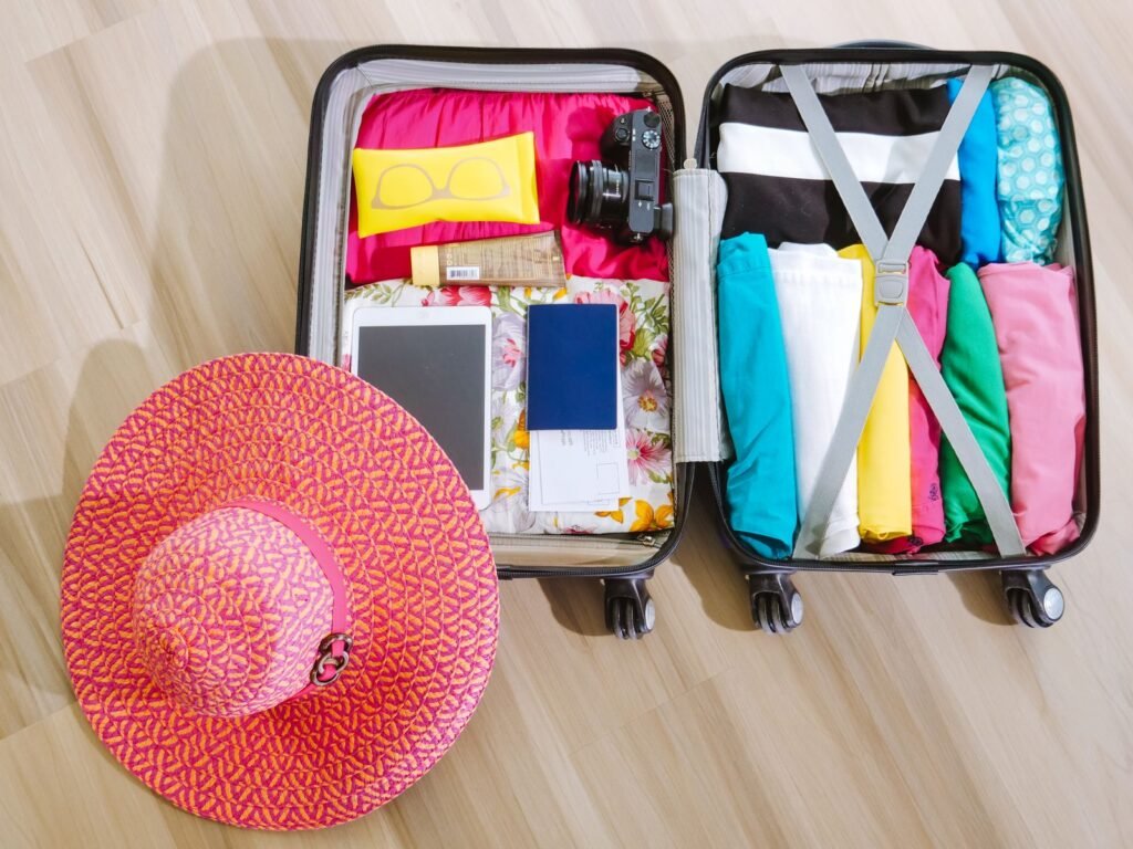 Top view of an open carry-on suitcase packed with rolled clothes, a camera, a tablet, a passport, and a sun hat