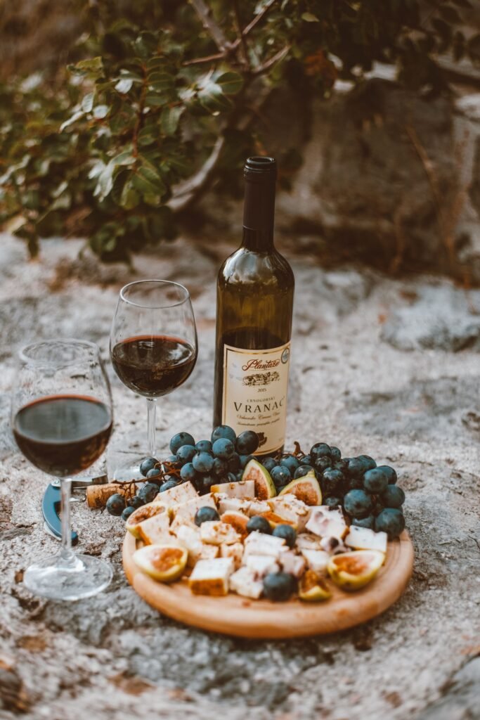 Two glasses of red wine beside a bottle, grapes, and a snack board outdoors