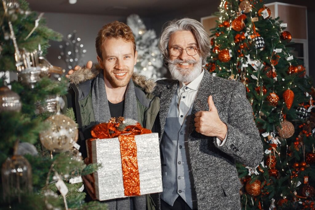 Adult son and his smiling father holding a wrapped Christmas gift by a decorated tree