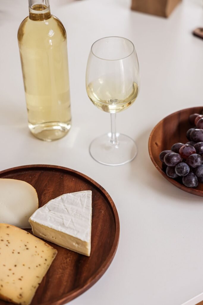 A glass of white wine beside a bottle, a cheese board, and grapes