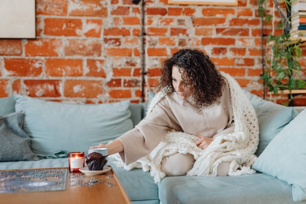 Woman wrapped in a knit blanket relaxing on a sofa with a warm drink and a candle
