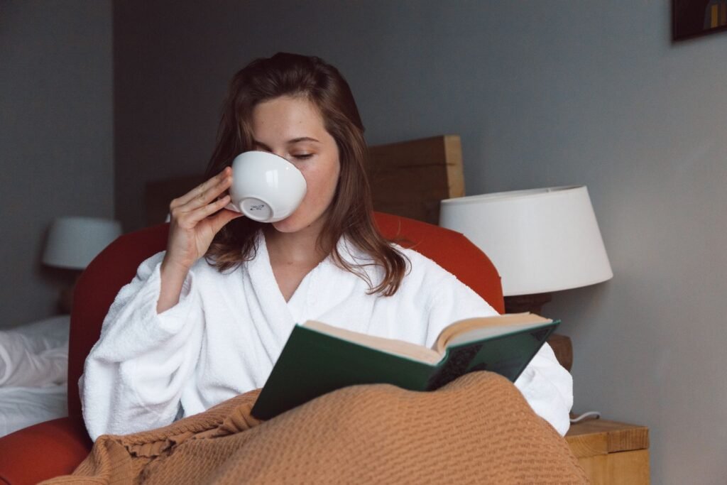 Woman in a white robe sipping coffee and reading a book under a blanket